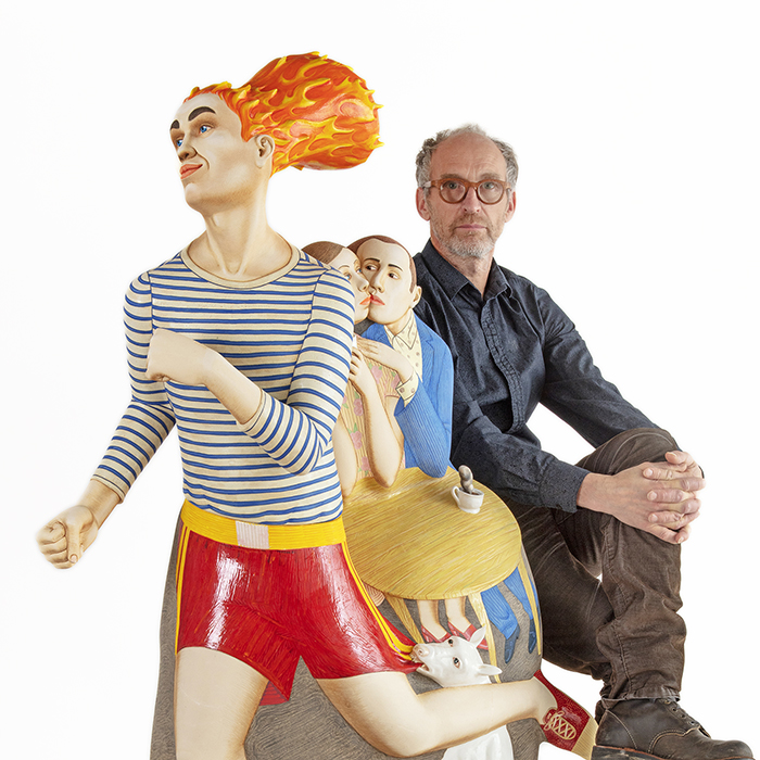 Sergei Isupov, "On the Way", 2020, porcelain, slip, glaze, 59.5 x 26 x 16". Portrait of the Artist, seated to the right, with arms crossed over one knee, which is resting on the other leg. He is seated behind his sculpture, On the Way, which is brightly colored in primary colors and patterns. The Central Figure is running away from a dog, which has hold of their red boxing-looking gym shorts. Behind this scene is a couple, seated at a paler yellow table, on the verge of a kiss or staring deeply into each others eyes. A single cup of coffee sits steaming on the table.