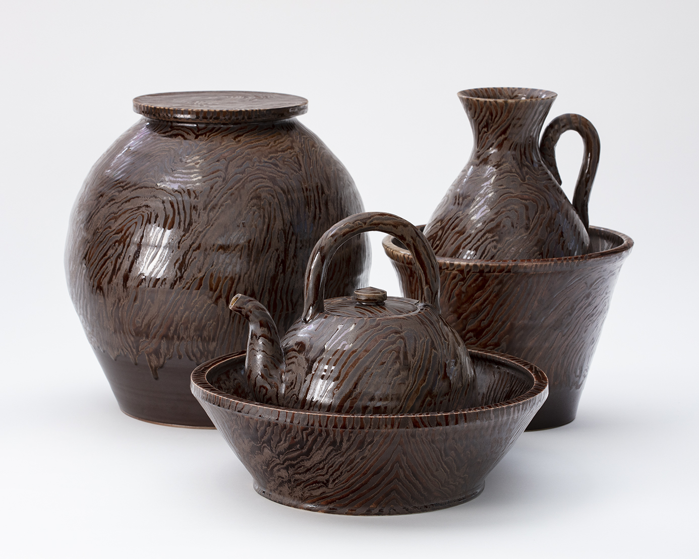 Linda Sikora, 'Faux Wood Group'. 2014-2018, stoneware, polychrome glaze, group footprint 12" x 3' x 2', Individual works: 5"-12" in height. Collection of the Smithsonian American Art Museum, 2021. John Polak Photography - group photo with large jar with flat lid, teapot within a wide basin, and handled pitchers within a taller and more narrow basin/bowl